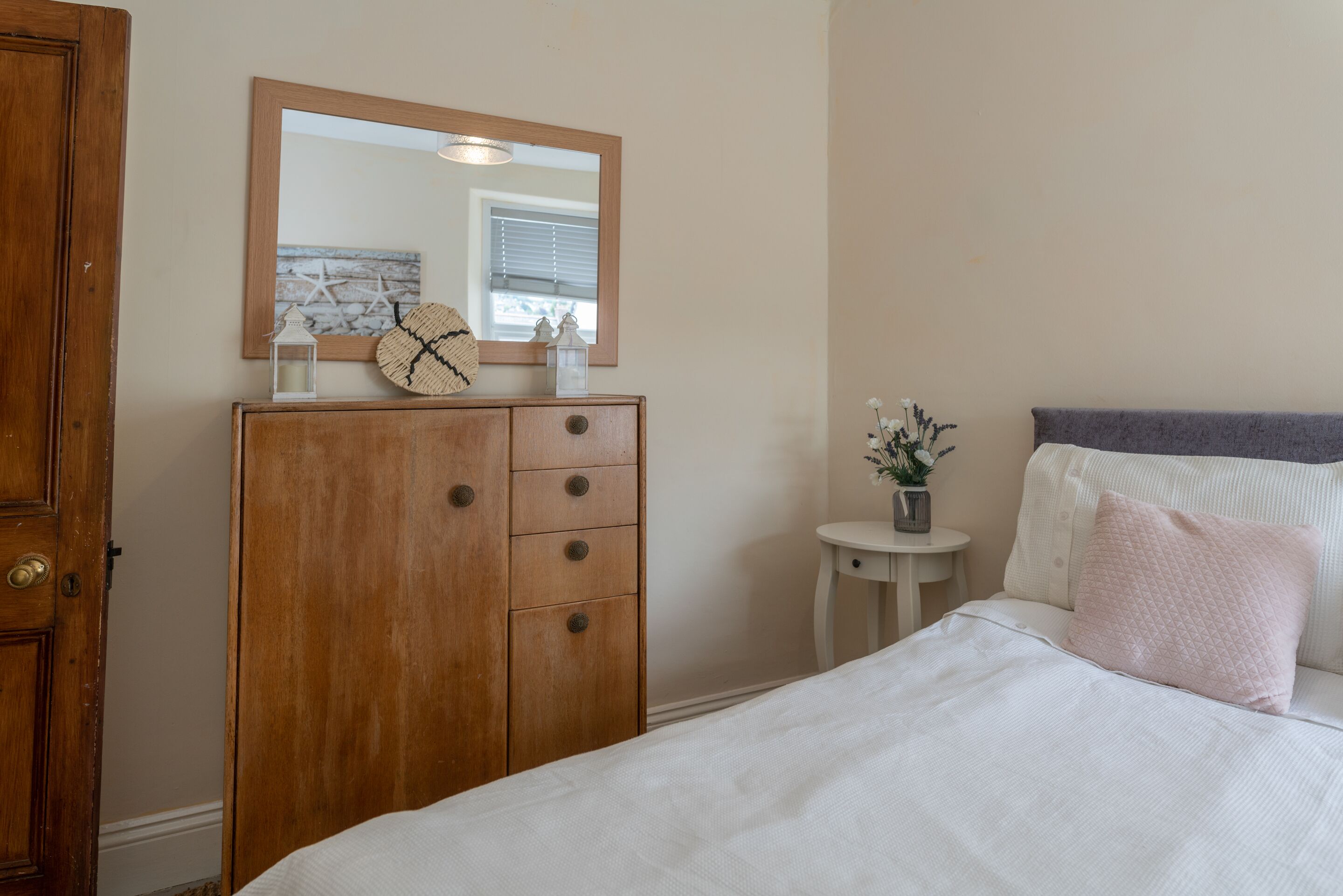 Une chambre avec une commode en bois, un miroir rond, une table de chevet ornée d’un vase de fleurs et un lit recouvert d’une couette blanche et agrémenté d’un coussin rose.
