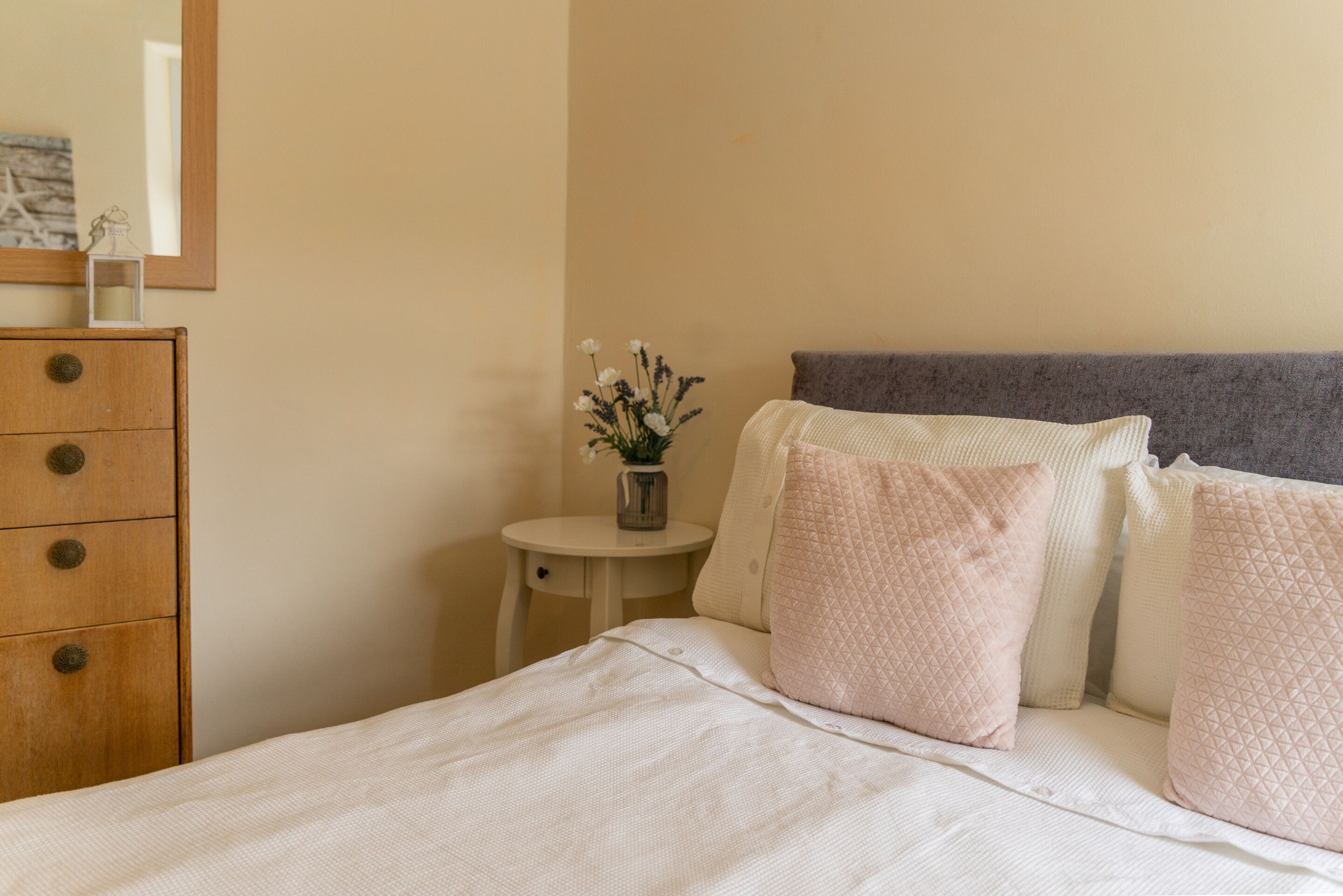 Een net opgemaakt bed met een grijs hoofdeinde, een houten dressoir met lades, en een tafeltje met een bloemenvaas.
