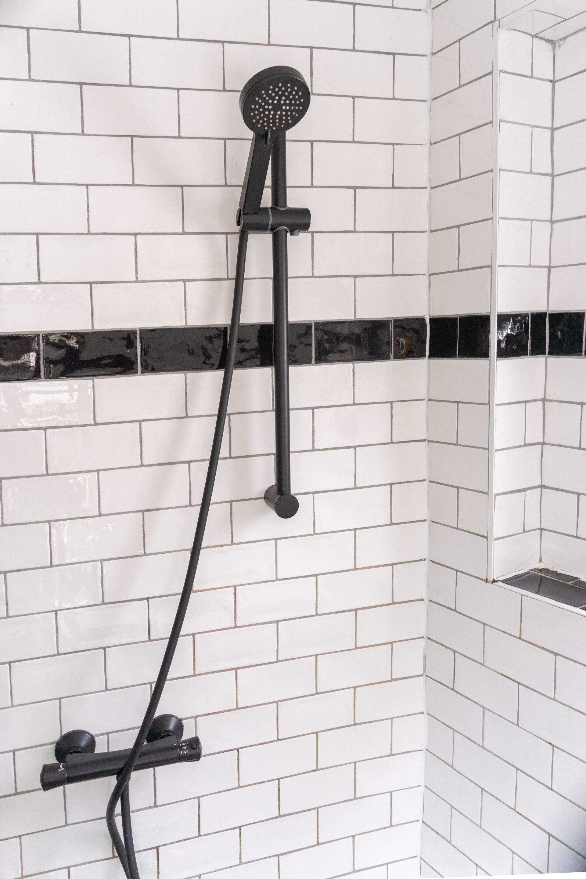 Una ducha con cabezal de ducha manual, montado sobre un soporte de metal negro, frente a una pared de azulejos blancos con líneas de lechada negra.
