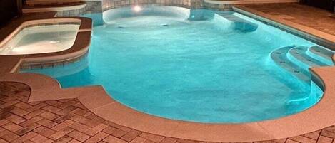 Piscina con un jacuzzi, rodeada por un patio de ladrillo y una zona cubierta con sillas.
