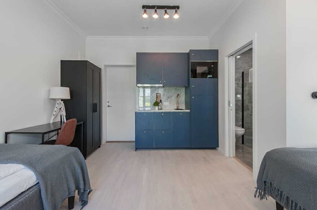 Una habitación pequeña con cama, escritorio, silla, kitchenette y baño.