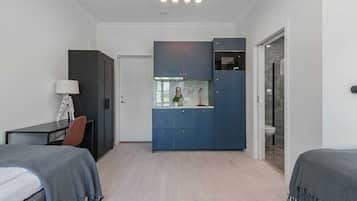 Una habitación pequeña con cama, escritorio, silla, kitchenette y baño.