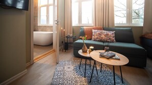 Una sala moderna con un ventanal grande, un sofá verde, una mesita redonda y una bañera en el baño contiguo.