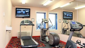 Gimnasio de hotel con cinta de correr, elíptica y bicicleta estática, un espejo grande y un televisor que muestra noticias.