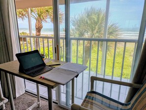 A laptop on a table with a notebook, a pen, and a mug, overlooking a balcony with a view of the ocean and palm trees.