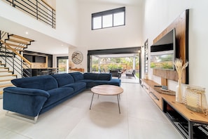 Ein modernes Wohnzimmer mit einem blauen Ecksofa, einem Holztisch, einem an der Wand befestigten Fernseher und einer Treppe, die zu einer oberen Ebene führt.