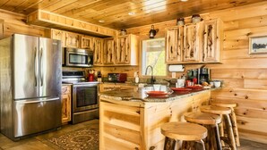Cocina de cabaña de madera con refrigerador, horno y microondas de acero inoxidable. Hay taburetes de madera en la isla y un suelo de madera rústico.