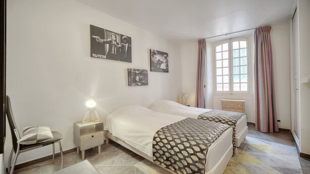 A bedroom with two beds, a chair, a bedside table with a lamp, and a window with curtains.