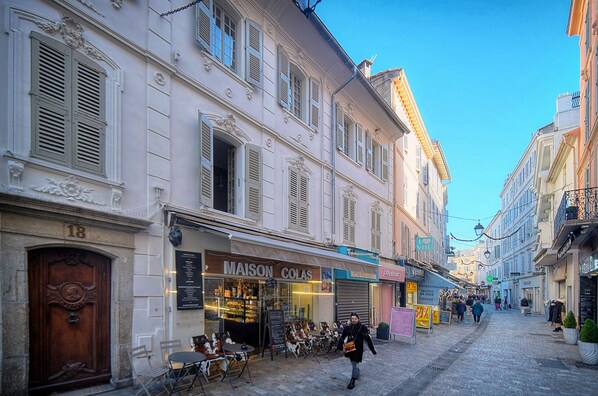A street with buildings, a Maison Colas shop, outdoor seating, and a person walking.
