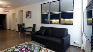 A modern living room with a black sofa, a glass coffee table with colorful rings, a dining area with chairs, and a kitchen visible through a window.