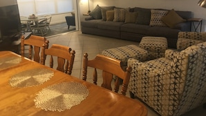 Una sala de estar con un sofá gris, otomanas estampadas, una mesa de comedor de madera con individuales y un televisor en una esquina.
