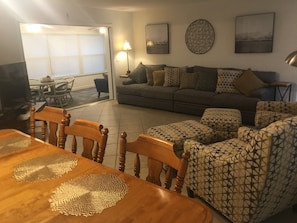 A living room with a grey sofa, patterned ottomans, a wooden dining table with placemats, and a TV in the corner.