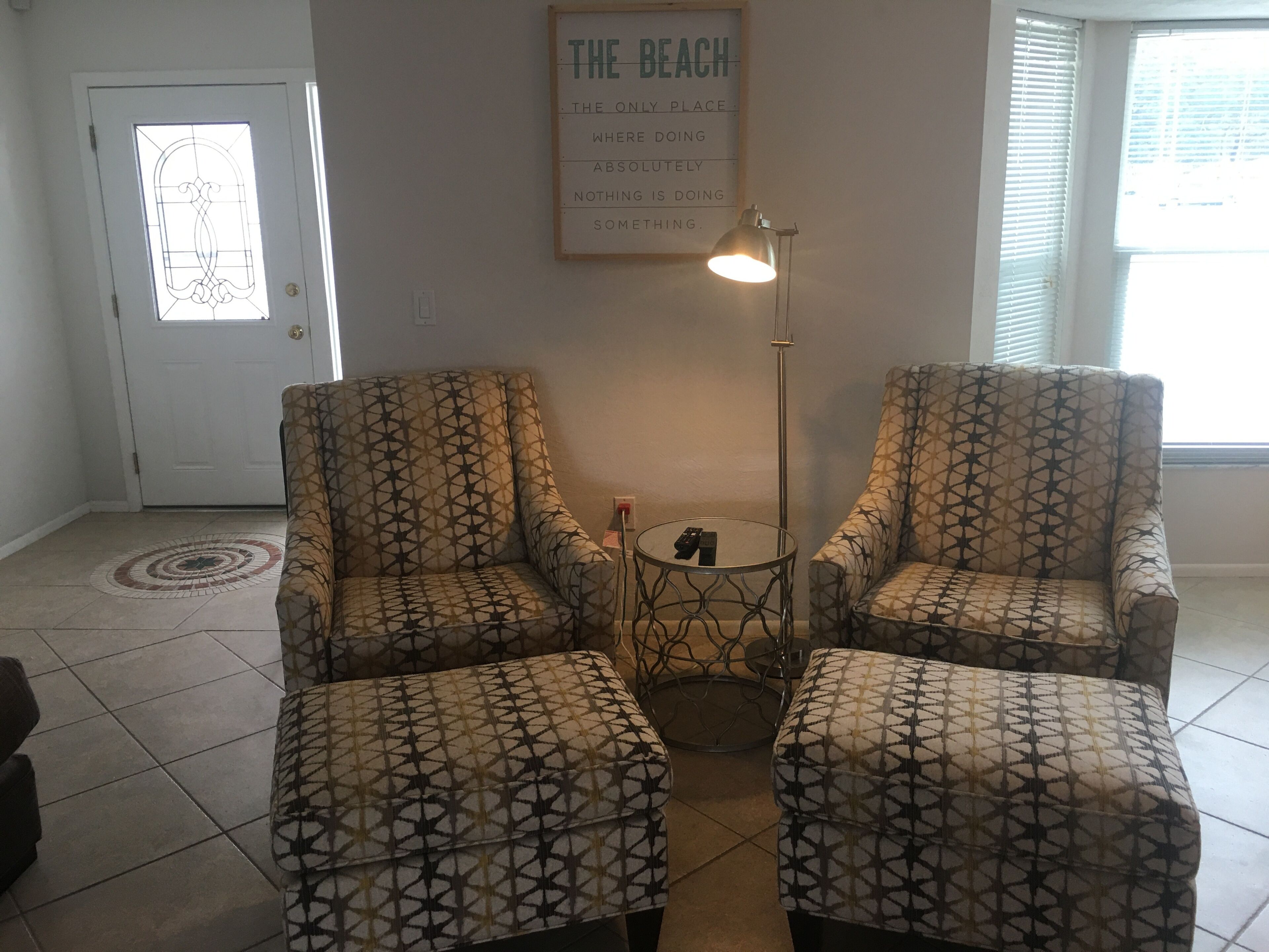 A living room with two patterned armchairs and ottomans, a small round table, a floor lamp, and a poster on the wall.