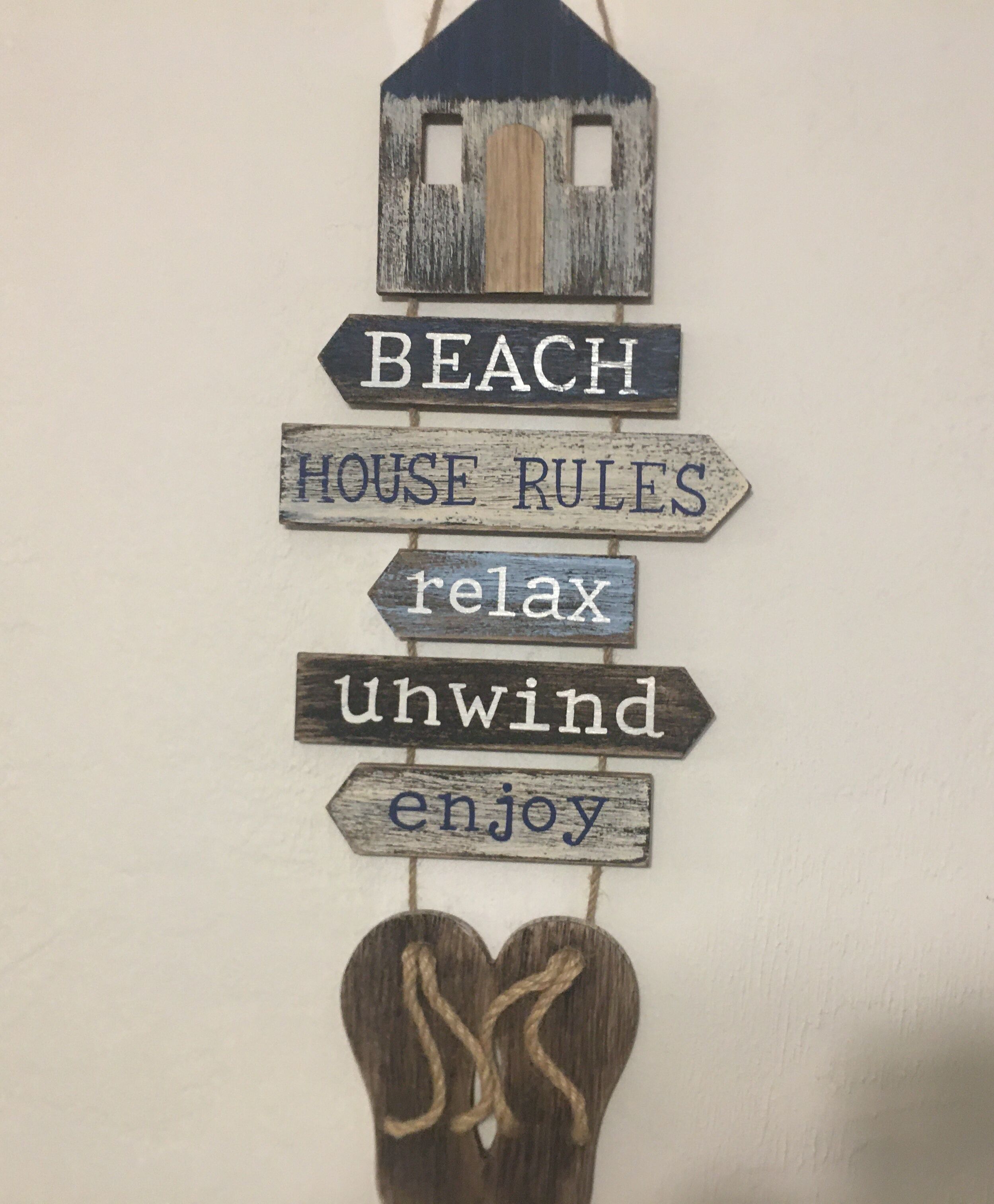 Eine Wanddekoration mit Holzschildern, auf denen steht: 'BEACH HOUSE RULES relax unwind enjoy', und einer kleinen Häuserfigur.