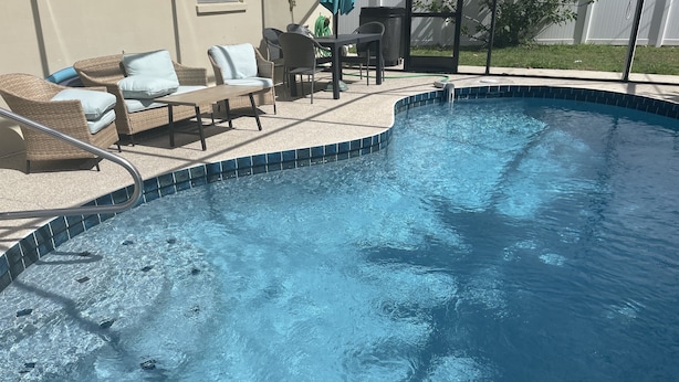 Una piscina con borde de azulejos, rodeada de mobiliario de mimbre y una mesa.