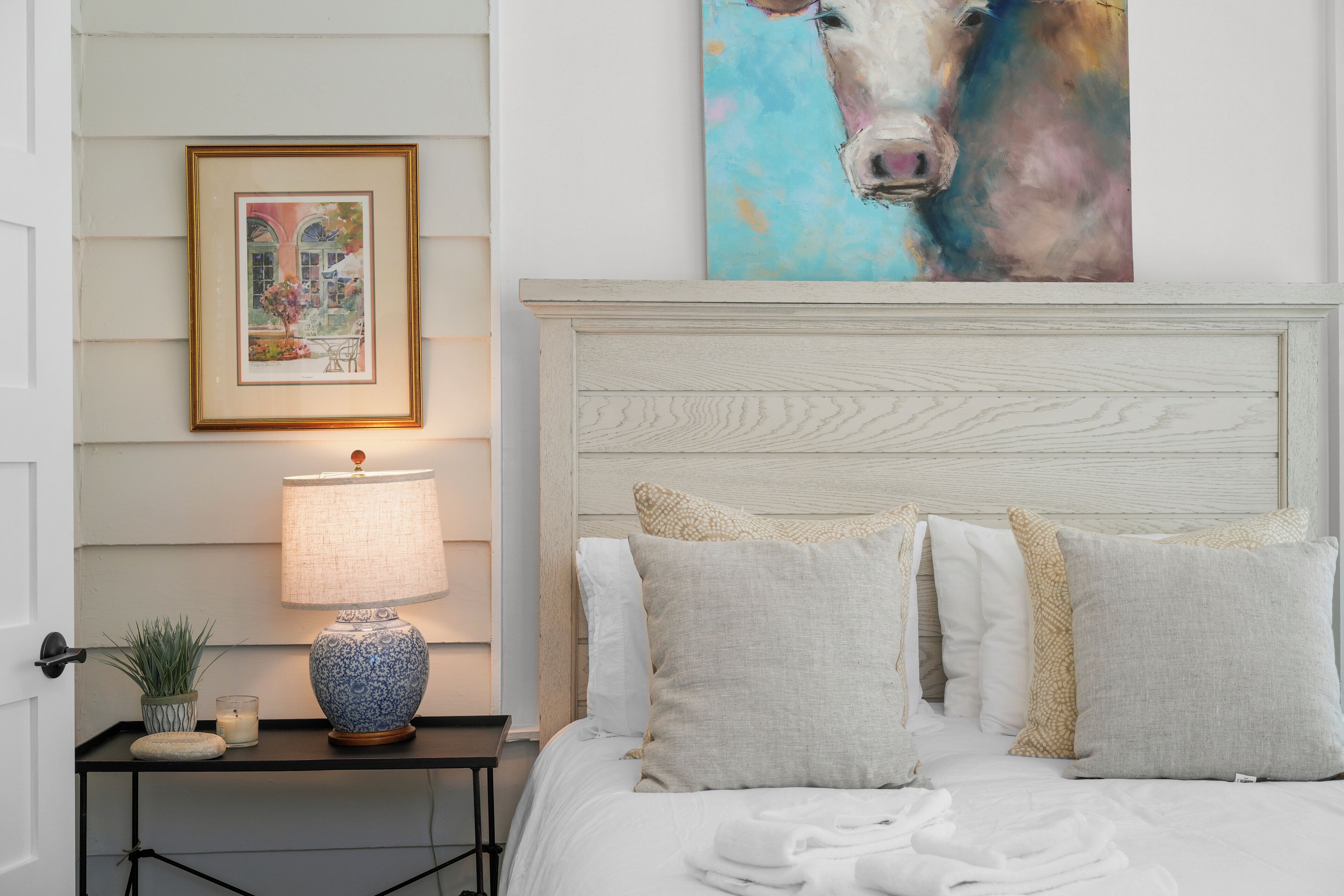 Een net opgemaakt bed met hoofdeinde, een nachtkastje met een lamp, een lijst met foto en een schilderij aan de muur.