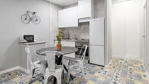 Een kleine keuken met witte kasten, een magnetron, een cactus in een pot en een eethoek met stoelen.