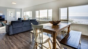 A spacious living room with a large window overlooking the ocean, a wooden dining table, and a gray sofa. - Ocean Front with Spectacular Views! 2 Bedroom, 2 Bath! Seal Rock! (Seal Rock)