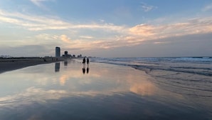 Una playa con arena húmeda que refleja el cielo, algunas personas caminando y edificios a lo lejos.