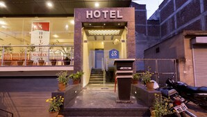 Entrada de hotel con un letrero que dice “HOTEL” sobre la puerta, un mostrador de recepción con un cartel de “BIENVENIDOS” y plantas en macetas en los escalones.