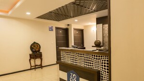 Zona de recepción de hotel con mostrador, una pequeña estatua en una mesita auxiliar y un cartel con el número de la habitación.