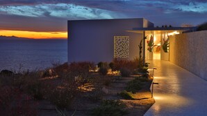 A modern building with a glass facade, a pathway leading to the entrance, and a view of the sea at sunset. - Luxurious Antiparos Villa with Infinity Pool and Stunning Sea and Sunset views (Antiparos)