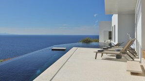 An infinity pool with a view of the ocean, lounge chairs, and a modern building facade. - Luxurious Antiparos Villa with Infinity Pool and Stunning Sea and Sunset views (Antiparos)