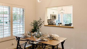 Un comedor con una mesa de madera dispuesta para cuatro personas, un jarrón con follaje y una cocina visible a través de una ventana sobre la mesa.