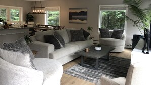 A spacious living room with a large gray sofa, a coffee table, and a dining area with a kitchen in the background. - The Mighty Oaks,Quiet,Hot Tub,Fire Pit,Cinema Room,Pool Table, 10 min to Town (Bar Harbor)