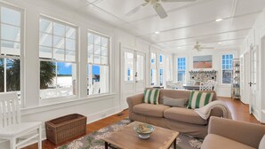 A spacious living room with a beige sofa, a wooden coffee table, and a white chair. There are large windows with white frames, a ceiling fan, and a view of the beach. - Back River Views, Wraparound Porch, Dock (Tybee Island)