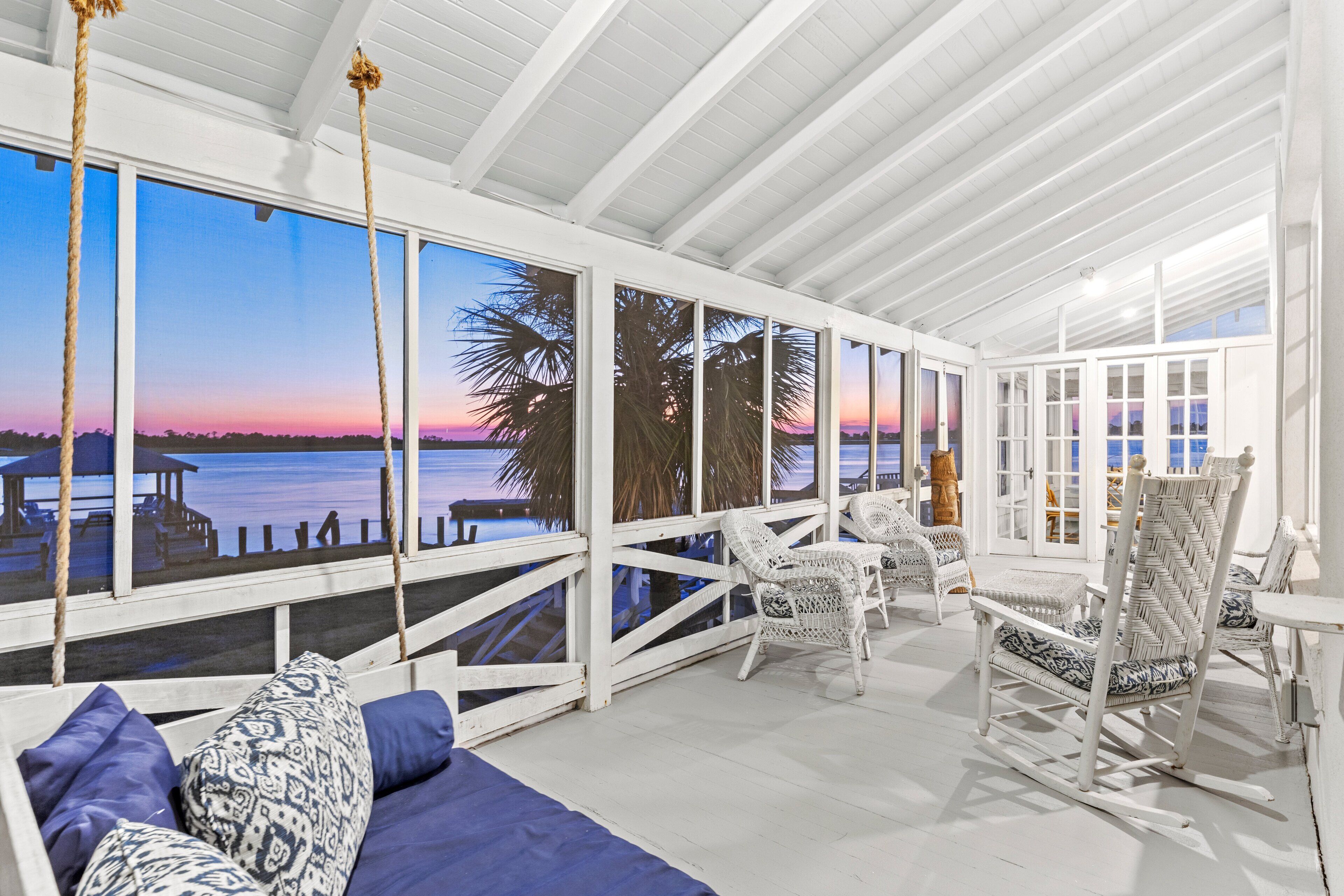 A covered outdoor seating area with white wicker furniture, a swing, and a view of the water at sunset.