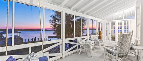 A covered outdoor seating area with white wicker furniture, a swing, and a view of the water at sunset.