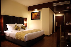 A hotel room with a large bed, a painting on the wall, and a desk with a chair. - Hotel Cama (Mohali)