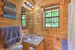 A cabin-style bathroom with wooden walls, a sink, a mirror, and a window with a view of greenery. - Quaint Log Cabin w/ Views Near West Jefferson (Warrensville)