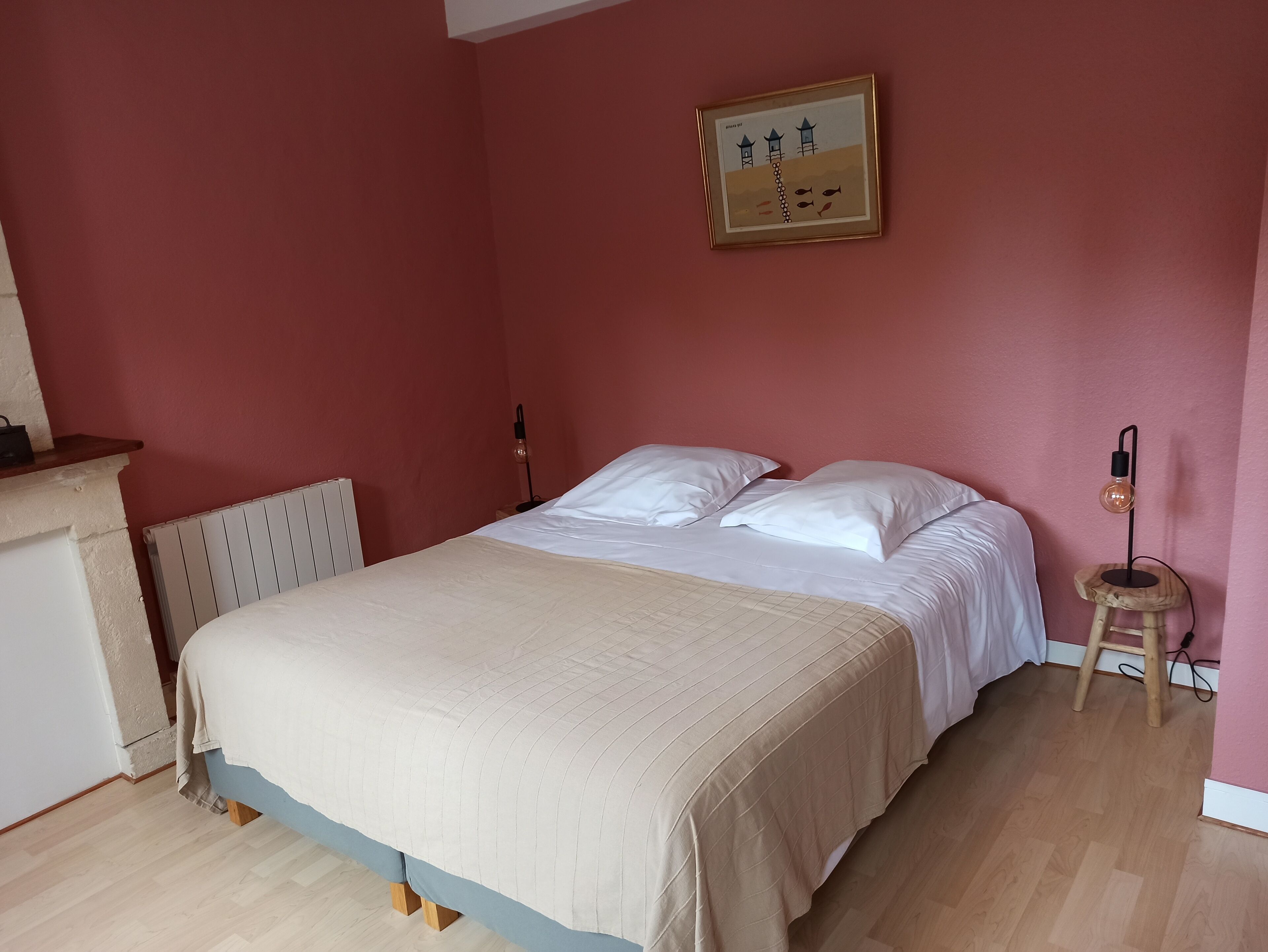 Une chambre à coucher comprenant un lit, une commode, un radiateur, un tableau encadré accroché au mur et un petit tabouret sur lequel est posée une lampe.