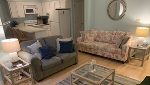 A living room with a grey sofa, a patterned sofa, a glass coffee table, and a kitchen area with white cabinets and appliances.