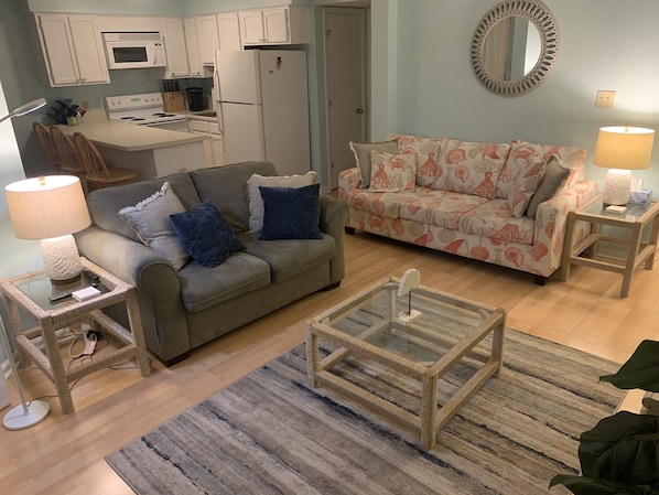 Sala de estar com sofá cinzento, um sofá estampado, mesa de centro de vidro e área de cozinha com armários e eletrodomésticos brancos.