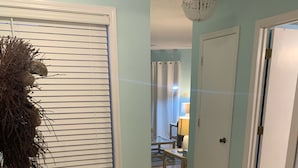 A hallway with light blue walls, a wooden floor, a wicker chest, and a door leading to a room with a lamp and a table.
