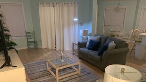 A living room with a glass coffee table, a sofa with cushions, a lamp, and a dining area with chairs.