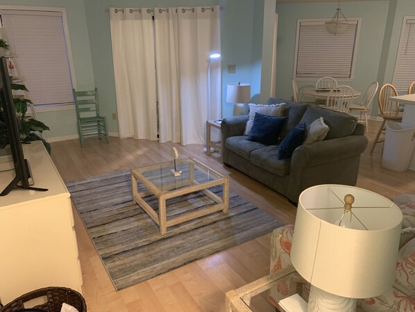 A living room with a glass coffee table, a sofa with cushions, a lamp, and a dining area with chairs.