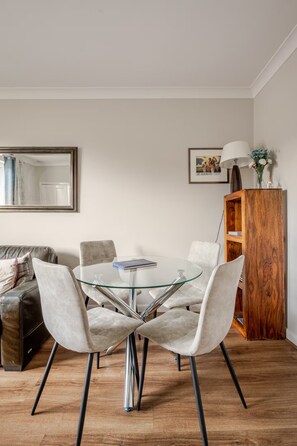 A dining area with a glass-top table surrounded by four chairs, a wooden cabinet, and a framed picture on the wall. - Central St Andrews Apartment | The Open 2027 | Walk to Old Course (St Andrews)