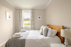 A hotel room with a bed, a nightstand, a lamp, and a window with curtains. - Central St Andrews Apartment | The Open 2027 | Walk to Old Course (St Andrews)
