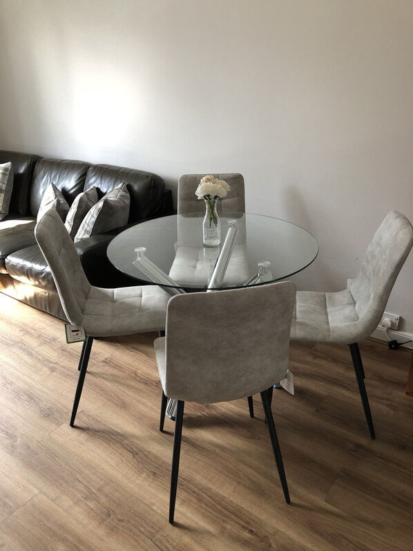 Un salon moderne avec une table à manger au plateau en verre, deux fauteuils gris à revêtement en tissu, un canapé en cuir noir et un vase avec des fleurs blanches.