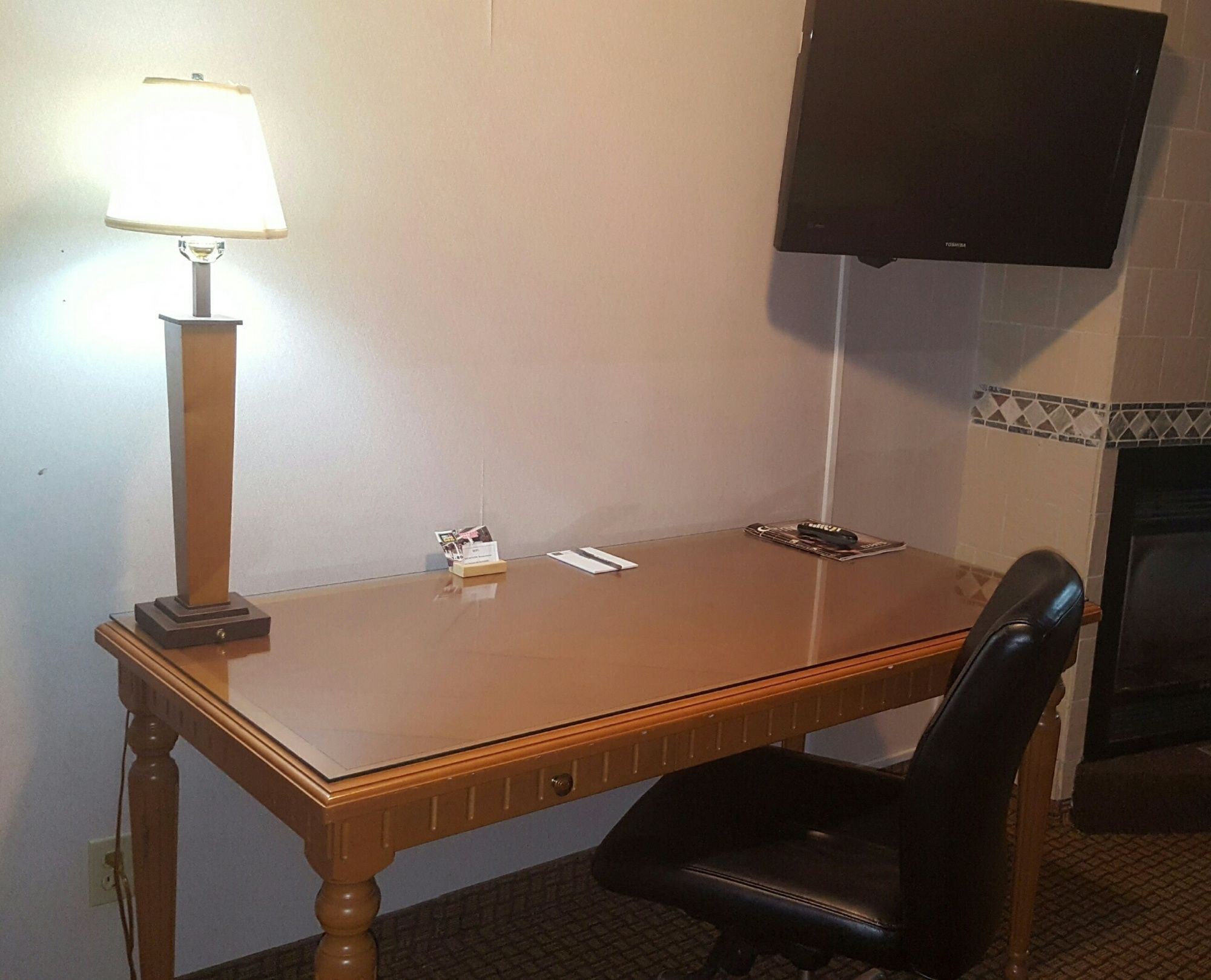 A hotel room with a wooden desk, a black leather chair, a lamp, a flat-screen TV mounted on the wall, and a small card stand with a card on it.