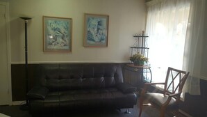 A living room with a black leather sofa, two framed paintings, a floor lamp, a wooden chair, and a small table with a vase of flowers. - OYO First Canada Hotel Cornwall Hwy 401 ON (Cornwall)