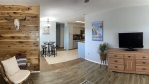 A modern living room with a wooden accent wall, a dining area with stools, a TV on a wooden cabinet, and a kitchen in the background. - Comfy, contemporary, quiet retreat with views of the Petroglyphs  (Albuquerque)