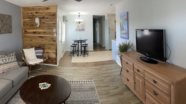 A living room with a ceiling fan, a wooden wall with a cow skull, a sofa, a coffee table, a TV on a wooden cabinet, and a dining area with stools. - Comfy, contemporary, quiet retreat with views of the Petroglyphs  (Albuquerque)