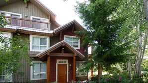 A two-story wooden building with a balcony on the upper floor, a front door, and a garden with shrubs and flowers.