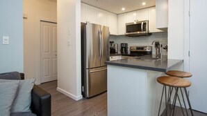 A modern kitchen with stainless steel appliances, a microwave, and a coffee maker. There are two wooden stools and a grey couch in the adjacent living area.