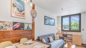 A modern living room with a dining area, wooden paneling, a clock, and a large window with a mountain view. - Appartement Courmayeur - Welkeys (Chamonix-Mont-Blanc)